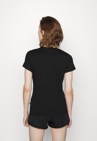 Mladá osoba s hnědými vlasy po ramena, oblečená v černém tričku s krátkým rukávem a černých šortkách, stojící na bílém pozadí.