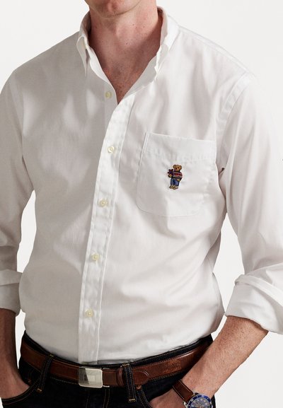 Vīrietis, kurš valkā baltu krekla ar pogām, uz kura krūšu kabatas ir izšūts lācītis, brūnu ādas jostu un zilas krāsas rokas pulksteni.