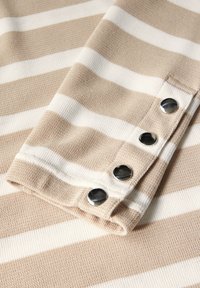 Beige und weiß gestreifter Stoff mit einer strukturierten Webart, der an einer Kante vier silberne Druckknöpfe aufweist und ein gefaltetes Manschetten-Aussehen erzeugt.