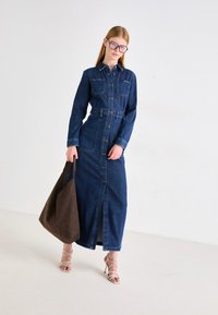 Vestido de camisa de denim, de manga larga, con botones en la parte delantera, dos bolsillos en el pecho, cintura ajustada, abertura hasta la rodilla, combinado con un tote marrón y tacones con tiras.