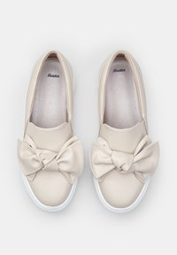 Beige slip-on kengät, jotka on valmistettu sileästä synteettisestä materiaalista, ja joissa on erottuva nauhakoriste sekä valkoiset kumipohjat.