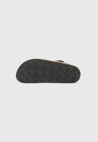 Semelle en sangle marron avec une semelle en caoutchouc noir texturé présentant un motif géométrique. Sangle de couleur claire pour un maintien sécurisé.