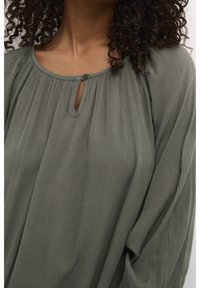Femme portant un chemisier ample à manches longues de couleur vert olive avec un col froncé et une petite fente avec bouton au niveau du col.
