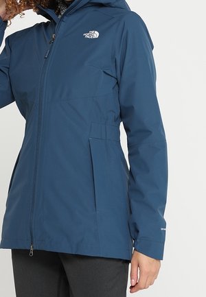 Person iført en blå North Face vandtæt jakke med lynlås foran, sidelommer og en hætte, vist fra brystet til hoften.