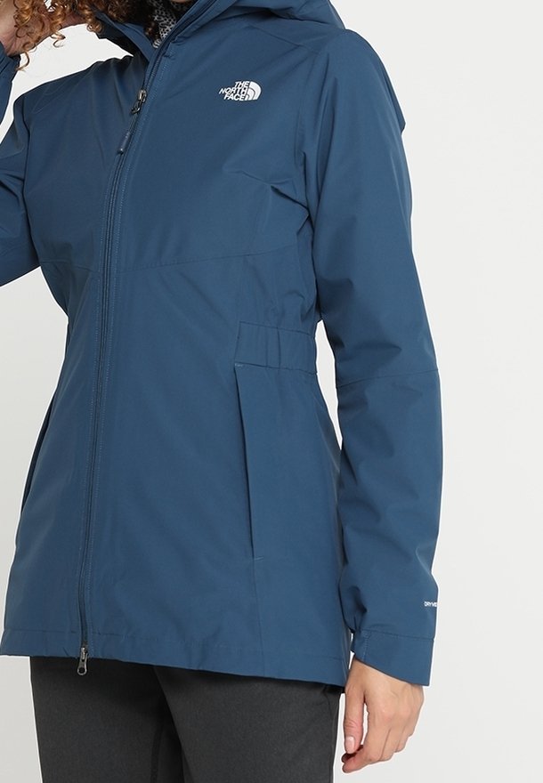 Personne portant une veste imperméable bleue North Face avec fermeture éclair devant, poches latérales et capuche, vue du buste aux hanches.