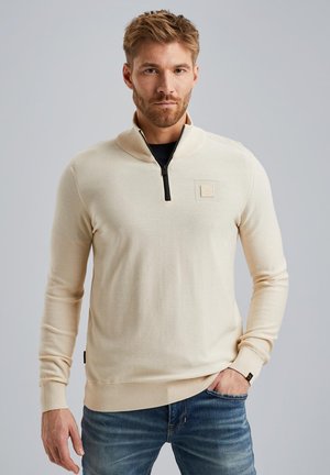 Beige zip-hals trui met een gestructureerde afwerking en ribgebreide manchetten, gecombineerd met blauwe jeans. Heeft een logo-patch op de borst.