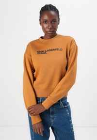 Mosterdkleurige sweatshirt met lange mouwen, geribde boorden en een ronde hals. Voorzien van een zwart "KARL LAGERFELD JEANS" logo aan de voorkant.