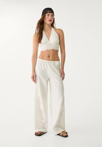 Hvid halterneck crop top med øjelet detaljer i kantene, parret med brede hvide bukser med blomsterudskæringer. Sort vævet pandebånd.