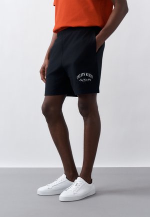 Persona con pantalones cortos negros Calvin Klein con logo blanco, camiseta roja y zapatillas blancas, de pie con una mano en el bolsillo contra un fondo claro.