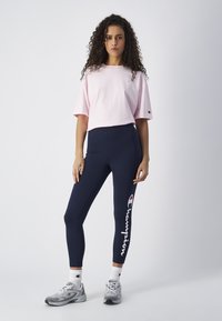 Jasnoróżowa bawełniana koszula z krótkim rękawem, granatowe legginsy z białym logo "Champion", szare trampki i białe skarpetki za kostkę. Jednolity tło.