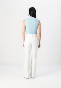 Woman with long dark hair wearing a sleeveless light blue top, white flared pants, and white sneakers, standing facing a white background.