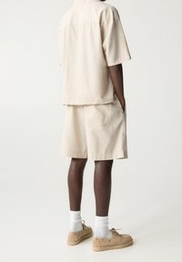 Chemise beige à manches courtes et shorts assortis en tissu léger. Chaussures marron clair avec une semelle texturée. Chaussettes blanches montant jusqu'à la cheville.