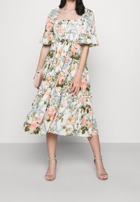 Vestido midi floral con escote cuadrado, mangas cortas con volantes, falda en capas, colores pastel y tejido ligero; combinado con un bolso de mano rosa y tacones.