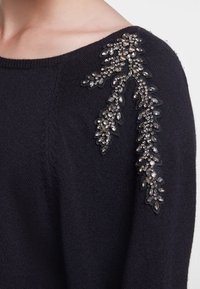 Pull noir avec une finition texturée, orné d'une embellissement floral en perles sur l'épaule gauche dans des tons argentés.