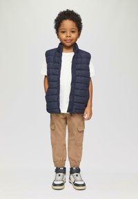 Marineblauwe gewatteerde vest over een witte shirt, gecombineerd met beige cargobroeken en hoge sneakers met marineblauwe en zilveren accenten.
