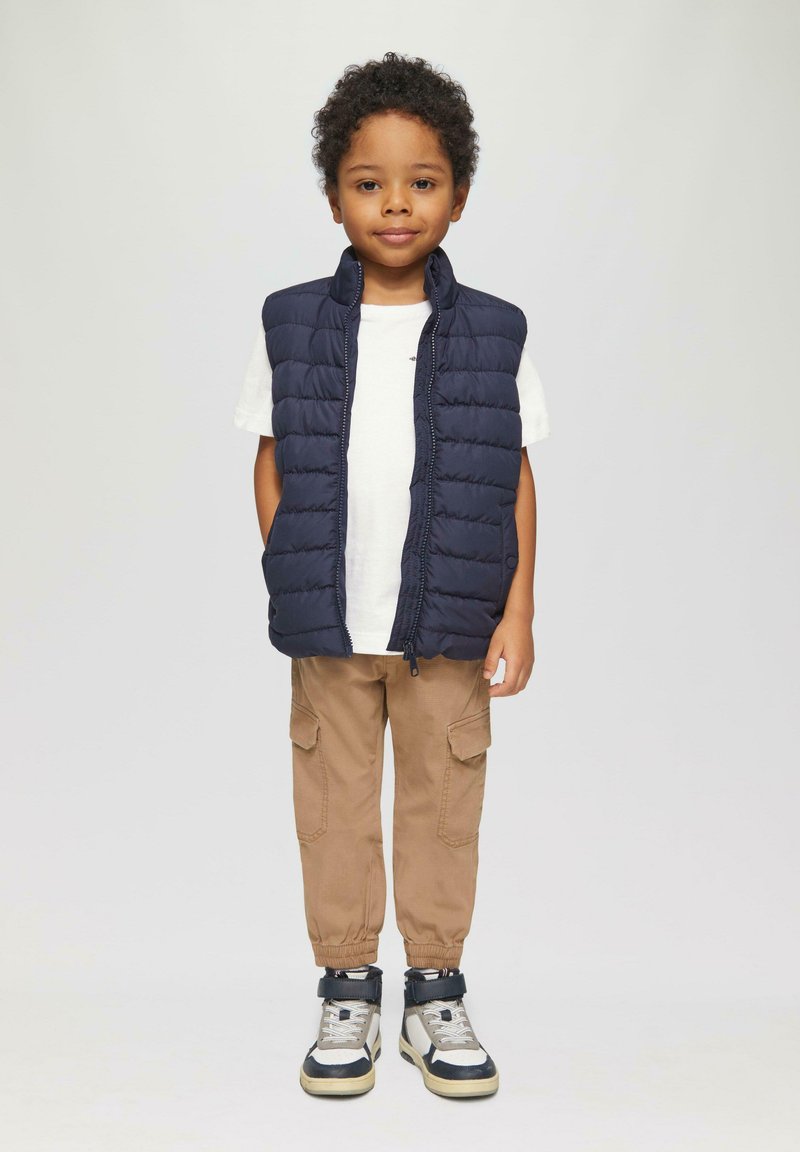 Marineblauwe gewatteerde vest over een witte shirt, gecombineerd met beige cargobroeken en hoge sneakers met marineblauwe en zilveren accenten.