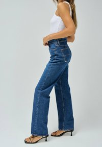 Jeans de mezclilla azul de cintura alta con corte recto, cinco bolsillos y acentos de costura. Combinados con una camiseta sin mangas blanca y tacones negros.