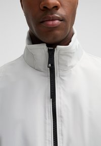 Helle graue Jacke mit hohem Kragen, aus einem glatten Material gefertigt. Verfügt über einen schwarzen Reißverschluss mit "STRELLSON"-Logo.