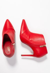 Botines de tacón alto de cuero sintético rojo con punteras puntiagudas, cremalleras laterales y una textura lisa. Altura del tacón aproximadamente 10 cm.