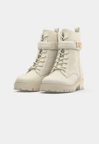 Beige Stiefeletten mit strukturierter Oberfläche, Schnürung vorne, verstellbarem Riemen, goldfarbenen Beschlägen und robustem Sohlen-Design.