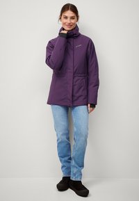Lila wasserdichte Jacke mit tailliertem Schnitt, Reißverschluss und Druckknopfverschlüssen sowie Kapuze. Kombiniert mit hellblauen Jeans und schwarzem Schuhwerk.