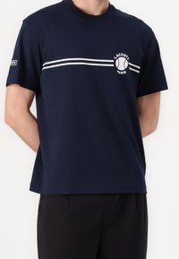Námořnická modrá bavlněná tričko s kulatým výstřihem, se bílými vodorovnými pruhy a kruhovým logem Lacoste Paris na hrudi.