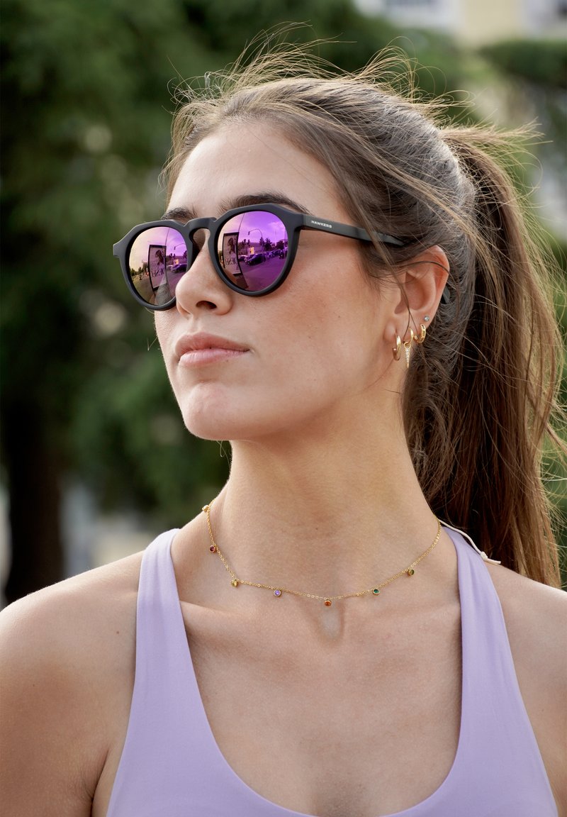 Fekete napszemüveg lila tükröződő lencsékkel, aranylánc színes díszítésekkel, és világoslila trikó egy modellen.