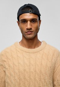 Beige kabelgebreide trui met een zachte textuur, gedragen met een zwarte baseballcap. Met een ronde halslijn en een losse pasvorm.