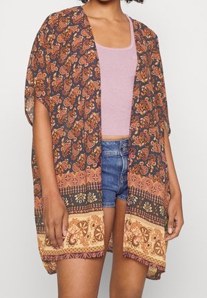 Kvinde iført blomsterprintet åbent kimono over en mauve top og denimshorts, stående mod en hvid baggrund.