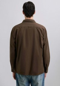 Bărbat cu părul închis la culoare, îmbrăcat într-o cămașă lungă maro și jeans albaștri deschis, stând cu spatele la un fundal simplu, deschis la culoare.