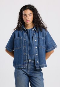 Giacca in denim a maniche corte di blu medio. Caratterizzata da bottoni a pressione, due tasche sul petto e una vestibilità rilassata con cuciture in evidenza.