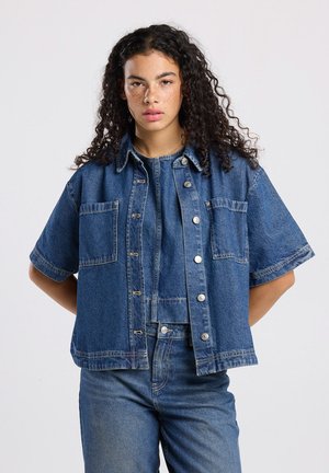 Kortere spijkerjack met korte mouwen in medium blauw. Bevat drukknoopsluitingen, twee borstzakken en een relaxte pasvorm met zichtbare stiksels.