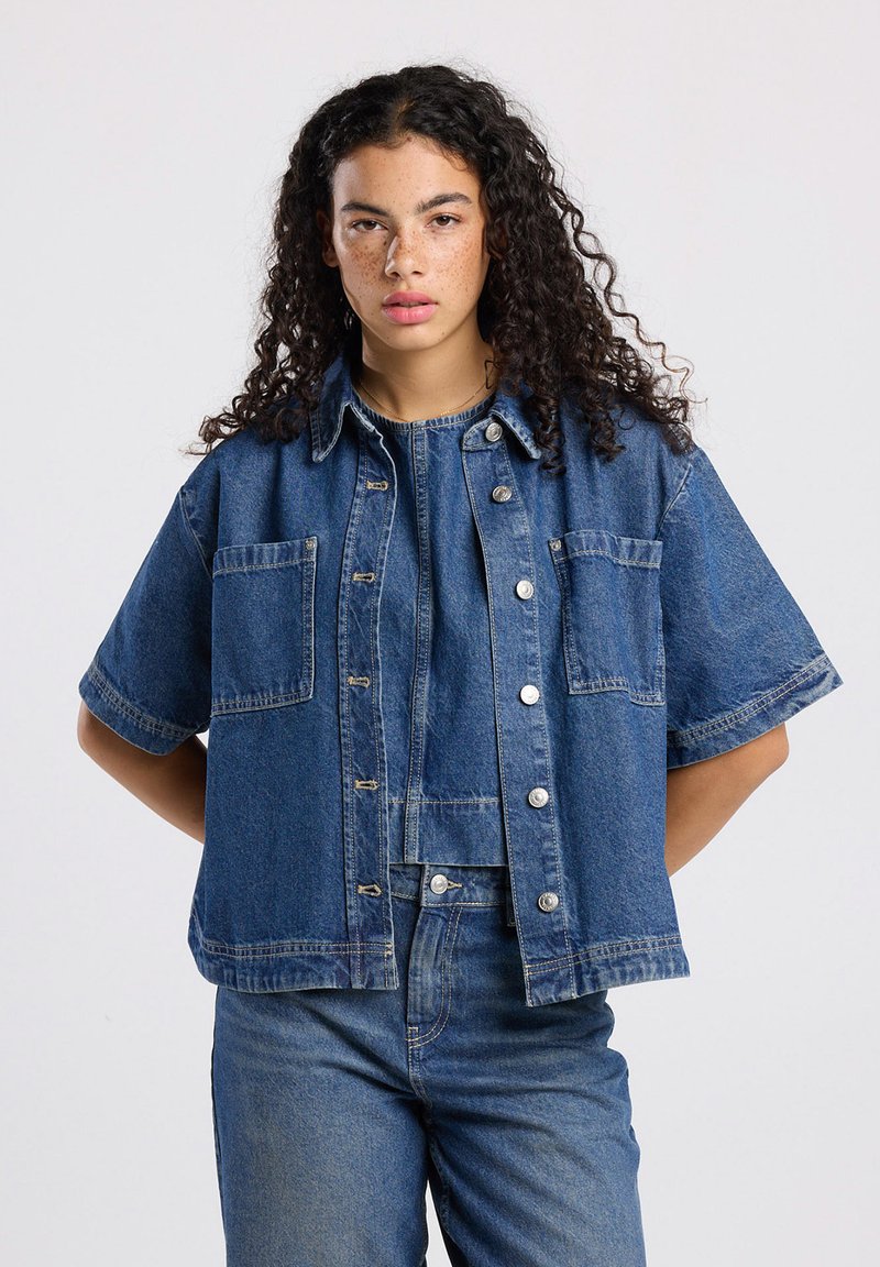 Giacca in denim a maniche corte di blu medio. Caratterizzata da bottoni a pressione, due tasche sul petto e una vestibilità rilassata con cuciture in evidenza.