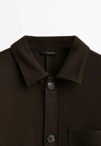 Chemise marron foncé à boutons avec col et coutures visibles, comprenant deux boutons noirs et une poche poitrine côté droit.