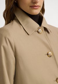 Beige kappa med uppvikt krage, utrustad med två runda knappar och en slät textur. Tyget verkar lätt och skräddarsytt.