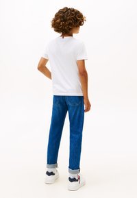 Kind mit lockigem Haar, trägt ein weißes T-Shirt, blaue hochgekrempelte Jeans, weiße Socken und weiße Turnschuhe, steht mit dem Rücken zum Betrachter.