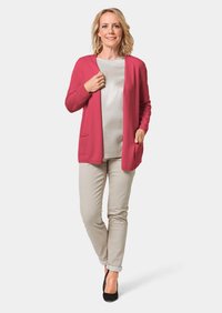 Roze cardigan met lange mouwen en een open voorkant, gedragen over een lichte top en gecombineerd met beige broeken en zwarte schoenen. Bevat twee zakken aan de voorkant.