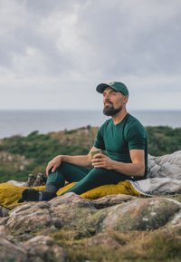 Vihreät urheiluvaatteet ja vihreä lippalakki, istumassa kallioilla keltaisen makuupussin kanssa, pitäen kahvimukia, merenrannan taustaa vasten.