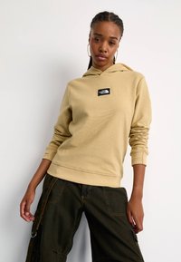 Beżowa bluza z kapturem o fakturowanej tkaninie, z czarną naszywką z logo. Żebrowane mankiety i dół. Połączona z ciemnymi spodniami cargo.