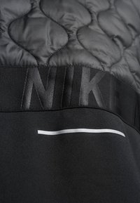 Gros plan d'un tissu matelassé noir avec le texte en relief « NIKE » et une fine bande horizontale réfléchissante sur un matériau noir lisse en dessous.