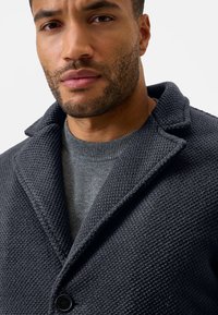 Kulgrå texturet frakke med sjalskrave og enkelt knaplukning, lagd over en grå sweater, der fremviser en glat stofoverflade.