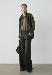 Veste en daim vert olive, t-shirt marron et pantalon à jambes larges gris foncé, avec un sac à main noir et une esthétique minimaliste.