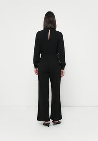 Zwarte jumpsuit van een gladde stof, met lange mouwen, een opening op de rug en een wijdvallend ontwerp, gecombineerd met zwarte hakken.