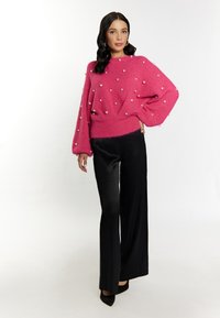Maglione a maglia rosa brillante con dettagli in perle, taglio oversize, orlo e polsini a coste, abbinato a pantaloni larghi neri e tacchi a punta.
