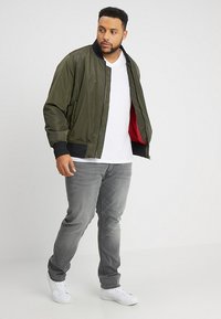 Giacca bomber verde oliva con colletto e polsini neri, caratterizzata da un interno rosso. Indossata sopra una t-shirt bianca e jeans grigi, con scarpe bianche.
