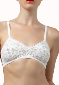 Donna che indossa un bralette bianco in pizzo floreale con spalline sottili, che mostra la parte superiore del torso e labbra rosse su sfondo bianco.