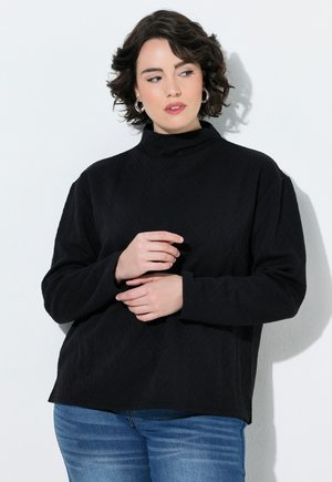 Ung kvinde med kort krøllet hår iført en sort langærmet top og blå jeans, stående mod en hvid væg, kigger til siden.
