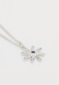 Pendente em forma de flor prateada com oito pétalas arredondadas e uma pequena pedra preciosa azul no centro, suspenso numa corrente de prata.