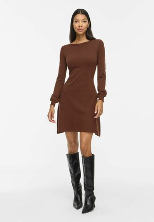 Femme portant une robe marron ajustée à manches longues et des bottes noires hautes à talons, debout devant un fond blanc.