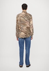 Svetla bež srajca z dolgimi rokavi in črnimi zebrastimi črtami; gladka tkanina; ima ovratnik in detajle z gumbi; v kombinaciji z modrimi kavbojkami.